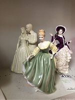 Front view of three porcelain figurines: Royal Doulton woman in green dress, seated woman in purple dress, and man and woman standing figurine.