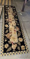 Full view of the rectangular rug runner placed on a concrete floor, showing the rooster and flower pattern with checkerboard borders.