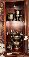 Tall china cabinet shelf with multiple silverplated serveware items including ice buckets, tea service, bowls, platter