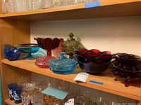Shelved display of various colored glass bowls and candle holders including red, blue, pink, and green pieces of varying shapes and sizes.