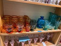Overview of amberina sherbet glasses, amberina tumblers, blue tumble-up, and teal glassware on wooden shelf