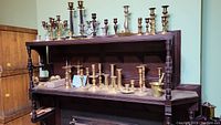 Full view of multiple brass candle holders of various sizes on two wooden shelves.