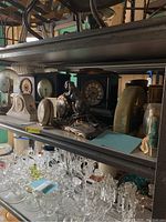Shelf with multiple vintage clocks and clock keys displayed among other glass items, showing various styles including a prominent black decorative clock with a feline figure.