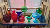Wide view of all colored glass items on wooden shelf, showing vases, decanters, and stemware in reds, greens, blues, and purples.