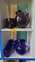 Amethyst glass pitcher with matching amethyst glasses on upper shelf and cobalt blue pitcher with matching cobalt blue glasses on lower shelf.