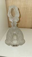 Full view of frosted glass decanter with detailed floral and leaf embossed pattern and large oval cutout stopper.