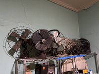 Shelf with seven fans including various vintage metal and one modern plastic fan