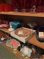 Overall shelf view showing cranberry goblets on top shelf and assorted Fenton pieces on lower shelf including aqua basket, small ruffled dishes and boxed carnival plate.