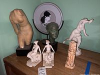 Overview of all items including nude torso sculpture with chips, green-toned bust, Fitz and Floyd bookends, nude fan-holding figurine, and decorative plate on stand.