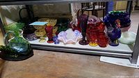 Assorted colored glass pieces on shelf including baskets, bowls, goblets and rooster figurine