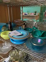 Wide view of multiple art glass bowls and candle holders on metal shelving, showcasing various colors including turquoise blue, cobalt, and clear glass pieces.
