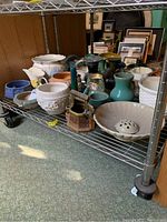 Wide shot showing multiple ceramic vases and pots on metal rack, varying colors and shapes, older vintage styles included