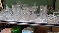 Shelf displaying multiple pressed glass items including pitchers, vases, bowls, and plates on one tier with green and brown dishes below.