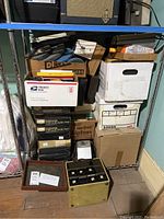 Stack of cardboard boxes including USPS box, labeled slide boxes, and metal film reels on floor under shelving