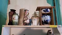 Overview showing five different sized ceramic and pottery vases and a stacked pair of white and blue floral patterned bowls on a shelf.
