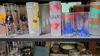Overview of 12 vintage beverage glasses with various World's Fair and vintage themes on a storage shelf