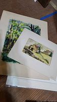 Three unframed signed watercolour paintings stacked on the floor showing rural scenes with barns and trees.