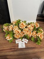 Overall view of the lot of artificial flowers showing many peach and white petaled flowers with attached green leaves laid on floor.