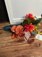 Lot of artificial flowers including orange and red flowers, green leaves, and stems on wooden floor
