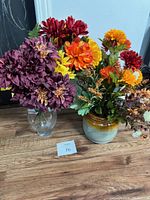 Two artificial flower arrangements in two vases placed on wooden surface