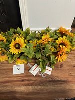 Lot of three brand new artificial sunflower stems with bright yellow flowers and green leaves, shown against wooden floor and white wall.