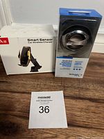 Both items in original packaging placed on wooden surface: smart sensor car wireless charger box (white) and SpeakerBall portable Bluetooth speaker in plastic package (silver sphere).