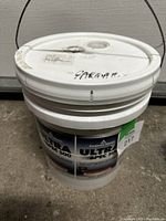 Full view of a large 5-gallon white plastic pail with handle and lid, labeled Benjamin Moore Ultra Spec 500 paint on concrete floor.