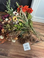 Lot of artificial flowers arranged on a wooden floor including orange, white, yellow, and green foliage with spiral brown accent pieces.