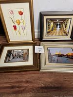 Photo showing four framed wall art pieces leaning against a wall on wooden floor, including tulip print, hallway, canal, and street scenes.
