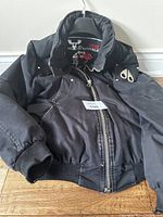 Front view of black Moose Knuckle jacket on hanger, showing zipper and sleeve patch.
