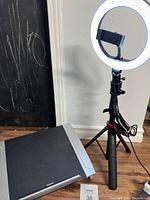 Short standing ring light on tripod with phone holder and power cord visible