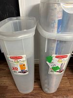 Two Rubbermaid Wrap Creator wrapping paper holders, one with wrapping paper rolls inside placed on wooden floor beside a wall.