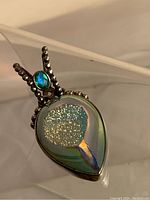 Close-up front view of the pendant showing polished blue iridescent teardrop geode with crystalline druzy and silver beaded bail with aquamarine accent.