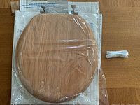 Natural wood toilet seat sealed in original plastic wrap on top of its packaging, showing the natural wood grain surface and attached hardware.