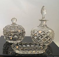 Oblong pin tray in front with lidded bowl and perfume bottle behind on black reflective surface, showcasing cut crystal design and clear glass details.