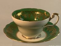 Side angled view of the green and gold decorated floral tea cup sitting on the matching saucer, showing detailed rim and cup shape.