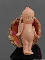 Front view of the small vintage celluloid Kewpie doll standing beside the colorful woven basket, showing facial features and body details.