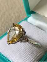 Pear shaped yellow tone gemstone on ring with silvertone band and smaller white stones to sides, displayed in a turquoise ring box.