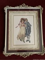 Full view of the framed needlework showing the Pink Girl and Blue Boy merged figures within the decorative frame with maroon velvet lining and gilt accents.