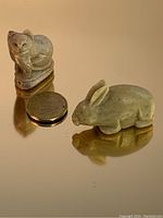 Photo showing two soapstone animal figurines, a cat and a rabbit, with a coin for size reference on reflective surface