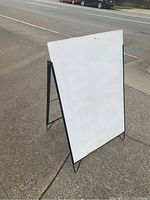 Metal framed folding sandwich board with white face, standing on a pavement with street in background.