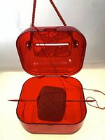 Open view of red celluloid yarn spool case showing inner spool and yarn passing through the slit in the lid.