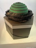 Front and side view of green fur felt cloche hat with black braided mohair spiral detail and trim atop original hatbox.