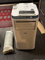 Front and top view of Arctic King portable air conditioner with attached user manual, remote control placed on top, and flexible exhaust hose on the floor next to it.