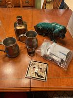 Photo showing pewter tankards, Blue Mountain Pottery green dog figurine, light switch cover, and picnic spoons in plastic bag on wooden table.