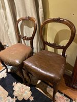 Pair of wooden backed chairs featuring carved wooden details and brown upholstered seats with wear, seen on floral rug and wooden floor.
