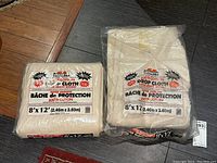 Two sealed packs of Bennett 100% cotton drop cloths, each measuring 8 by 12 feet, placed on floor near wooden trim.