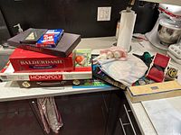 Overhead view of stacked board game boxes including Monopoly, Balderdash, Scrabble, and various accessories on kitchen counter