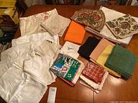Overview of folded assorted linens including embroidered tablecloths, napkins, and tea towels in multiple colors on wooden table