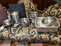 Overview of lot including variety of stainless steel pots, bowls, strainers, roasting pan arranged on patterned sofa